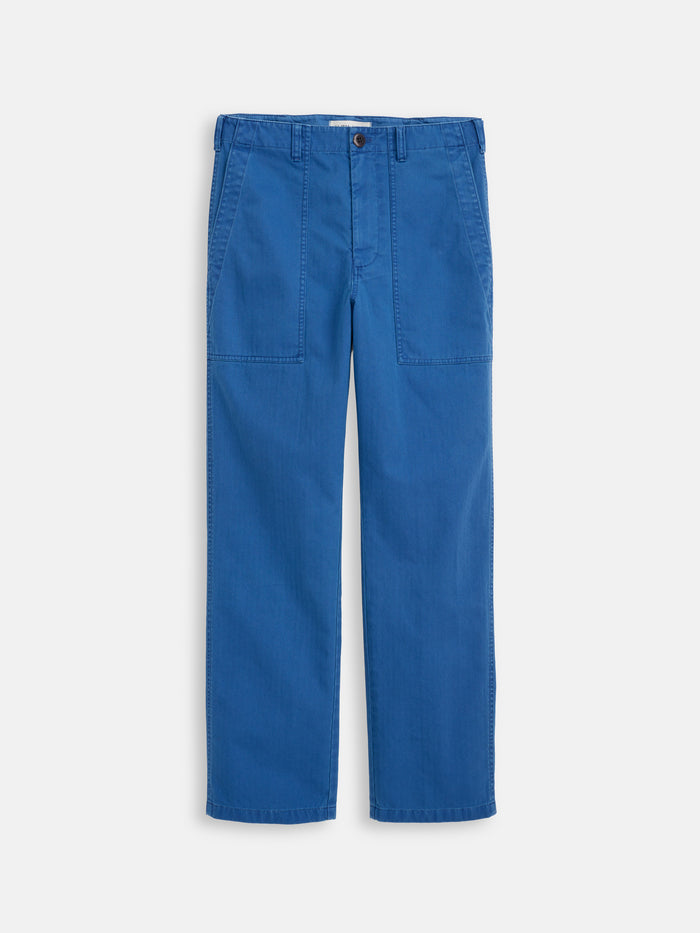 Neil Pant In Herringbone: bright blue straight-leg pants crafted from cotton herringbone with a garment-dyed finish, button and zip closure, two large front patch pockets, belt loops, and visible stitching. Shown laid flat on a white background.