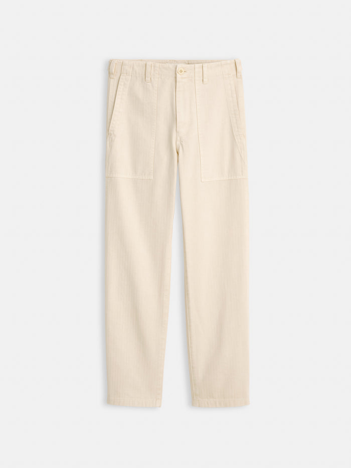 The Neil Pant In Herringbone: Beige straight-leg, utilitarian style pants made from garment-dyed cotton herringbone with front patch pockets, belt loops, and a button closure, shown on a plain white background.