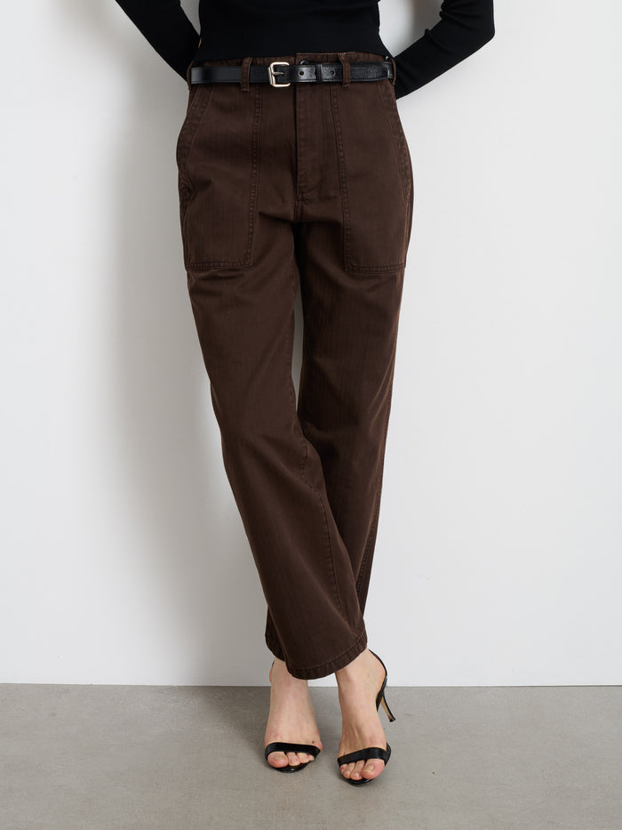 Someone wearing the Neil Pant In Herringbone, styled with a black belt, black long-sleeve top, and black open-toe heels, stands with legs crossed against a white wall and gray floor.