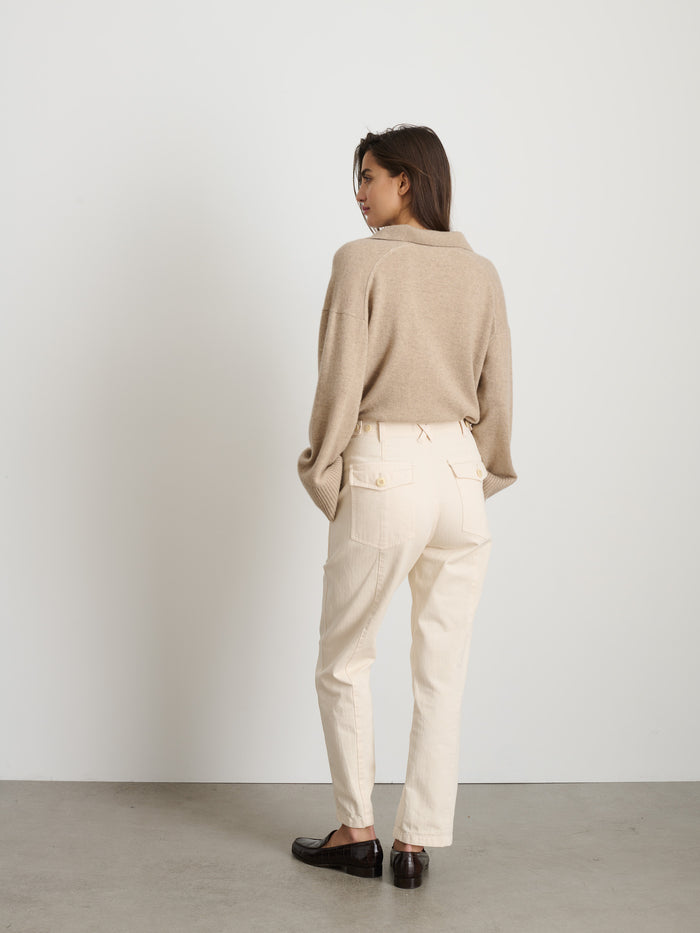 A woman stands indoors on a concrete floor with a white wall, wearing The Neil Pant In Herringbone—high-waisted cream cotton pants—paired with a loose beige sweater and dark loafers, creating an effortless utilitarian look.