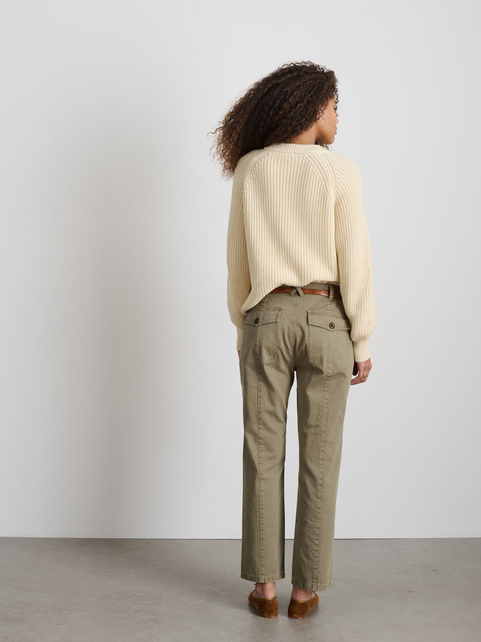 A woman with curly hair, facing away, wears The Neil Pant In Herringbone—olive green utilitarian style with back pockets—paired with a cream cotton sweater and brown shoes, against a white wall and gray floor.