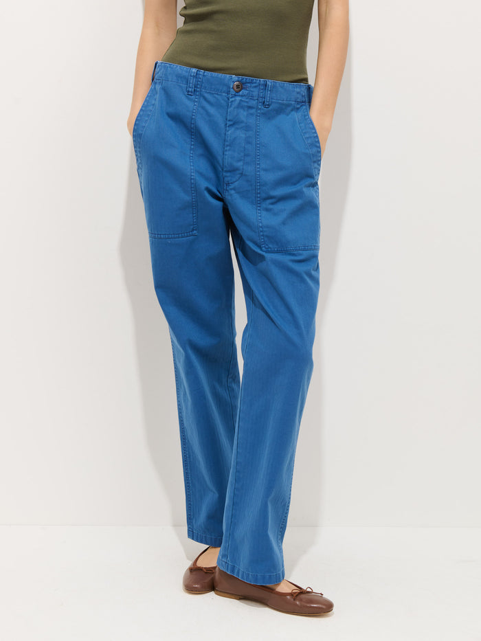 Someone stands against a plain white background, hands in pockets, wearing the Neil Pant In Herringbone—blue straight-leg cotton pants with front patch pockets—paired with a green top and brown flats for an effortless utilitarian look.
