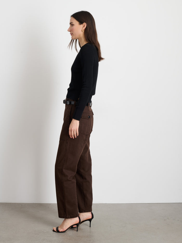 A woman with straight dark hair stands in profile against a white wall, wearing the Neil Pant In Herringbone, a black long-sleeve top, a black belt, and heeled sandals on a concrete floor.