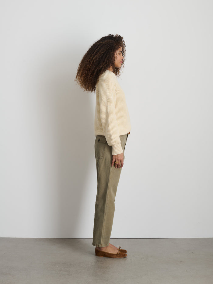 A woman with curly hair stands in profile against a white wall, wearing a garment-dyed cream sweater and The Neil Pant In Herringbone, paired with brown loafers for a relaxed utilitarian look, her arms resting at her sides.