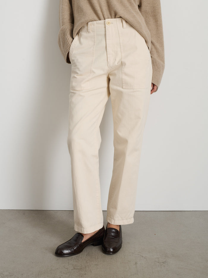 A person stands against a plain white wall and concrete floor wearing The Neil Pant In Herringbone in cream, a loose tan sweater with sleeves partly over their hands, and dark brown loafers.
