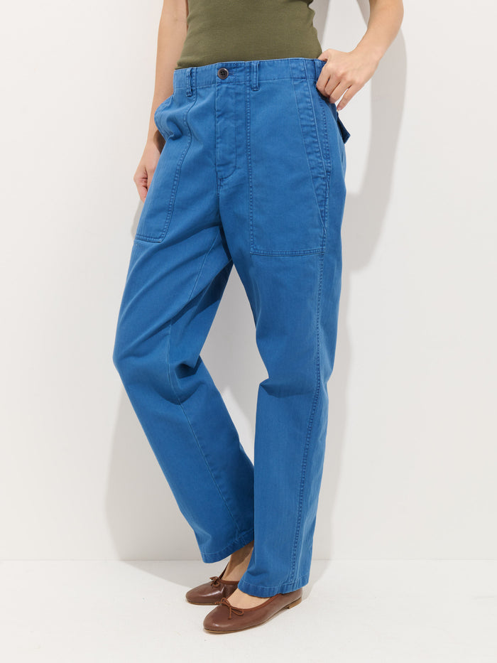 A person wearing the Neil Pant In Herringbone—loose-fitting, garment-dyed blue pants with large front pockets—paired with a green top and brown flat shoes, stands against a plain white background.