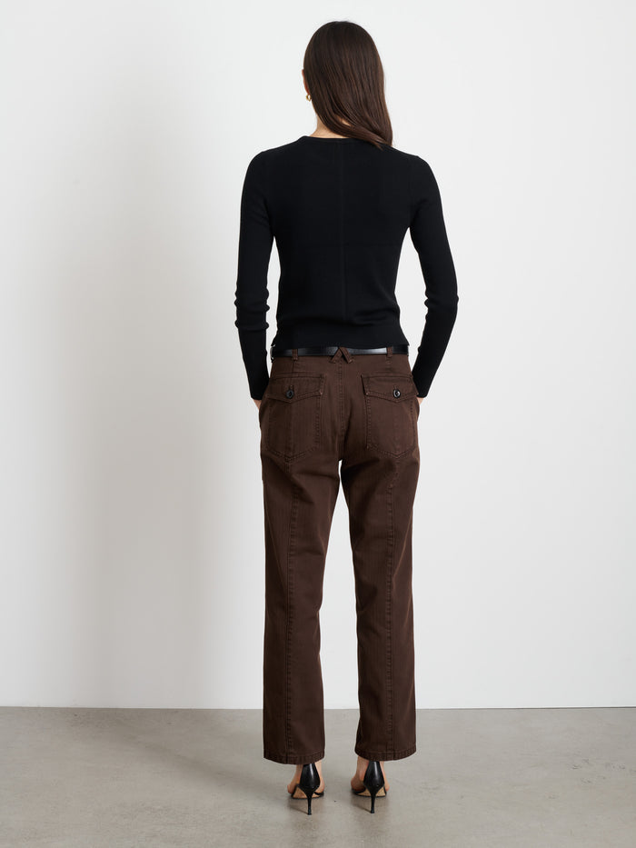 A woman faces away from the camera, wearing a black long-sleeve top, Neil Pant In Herringbone in brown, and black heels against a plain white wall and gray floor.