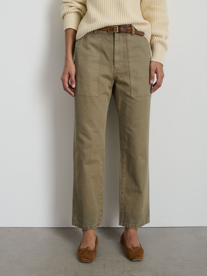 Someone stands against a white wall and grey floor in The Neil Pant In Herringbone—relaxed olive green wide-leg cotton pants with front patch pockets—styled with a brown belt, cream sweater, and brown moccasins for a utilitarian look.