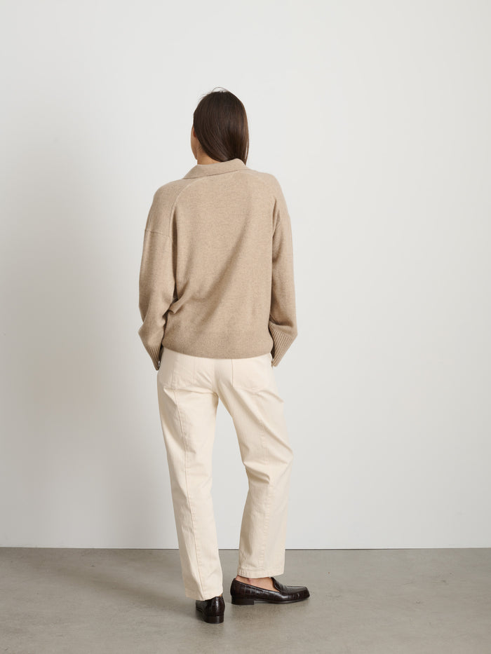 A person with straight, dark hair stands facing away, wearing The Neil Pant In Herringbone with a garment-dyed beige sweater and dark loafers, set against a plain white wall and concrete floor.