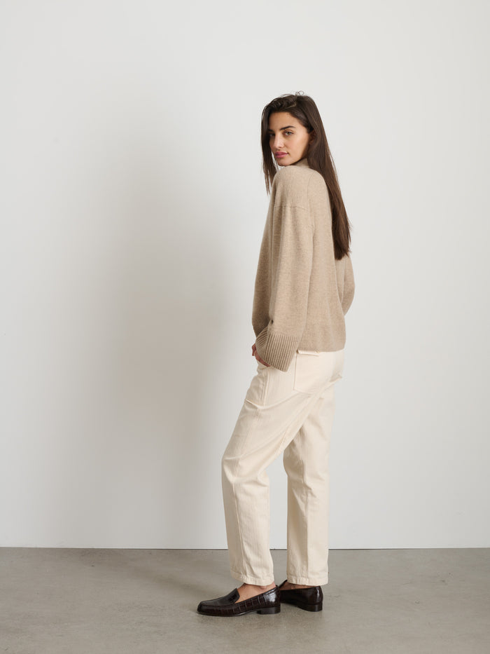 A woman with long brown hair, wearing The Neil Pant In Herringbone and a garment-dyed beige sweater, stands sideways in a minimalist studio with a plain white wall and concrete floor, glancing over her shoulder.