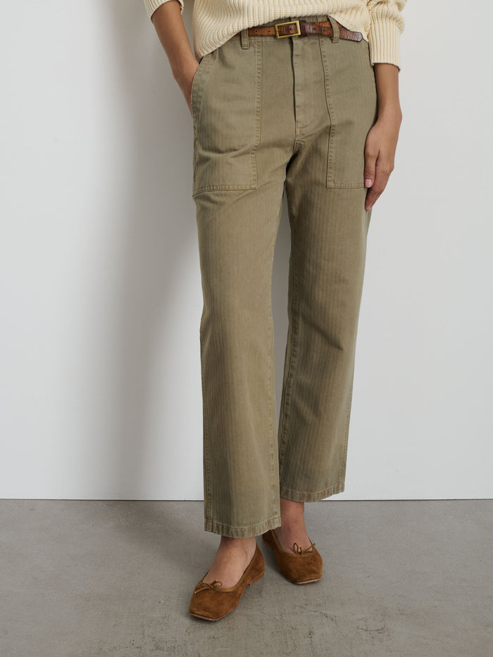 A person stands against a white wall wearing The Neil Pant in Herringbone—khaki wide-leg pants with front patch pockets—styled with a brown belt, cream sweater, and brown ballet flats. Their hands in the pockets add to the utilitarian look.