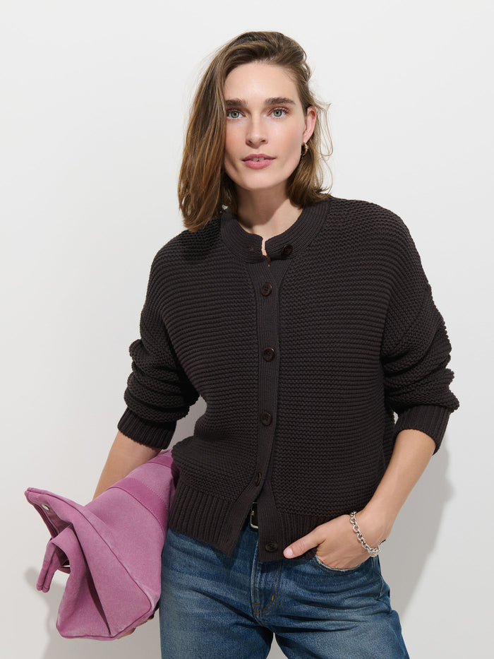 A person wearing the Nico Cardigan in Cotton—a textured dark women’s sweater with corozo buttons—paired with blue jeans stands against a white background, holding a folded pink coat in one hand.
