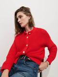 A person with shoulder-length brown hair wears the Nico Cardigan in Cotton—a textured red knit sweater with large corozo buttons—paired with blue jeans, seated on a chair against a plain white background to showcase a relaxed women's designer style.