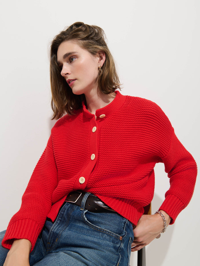 A person with shoulder-length brown hair wears the Nico Cardigan in Cotton—a textured red knit sweater with large corozo buttons—paired with blue jeans, seated on a chair against a plain white background to showcase a relaxed women's designer style.