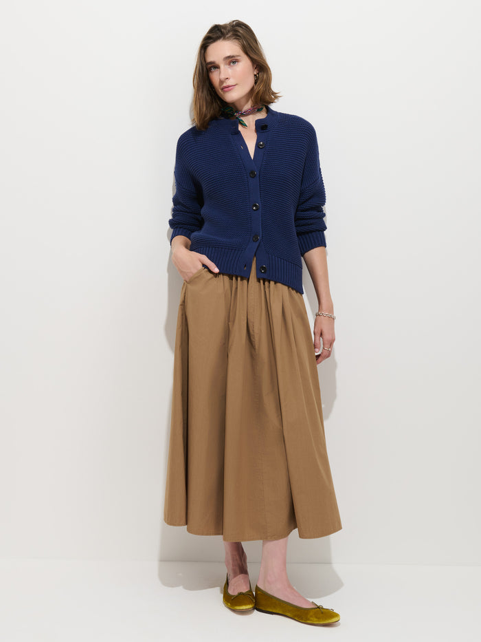 A woman stands against a white wall, wearing the Nico Cardigan in Cotton—a navy blue 100% cotton knit piece with Corozo buttons—paired with a tan midi skirt, yellow flats, and a small neck scarf. With one hand tucked into her skirt pocket, she gazes softly at the camera.