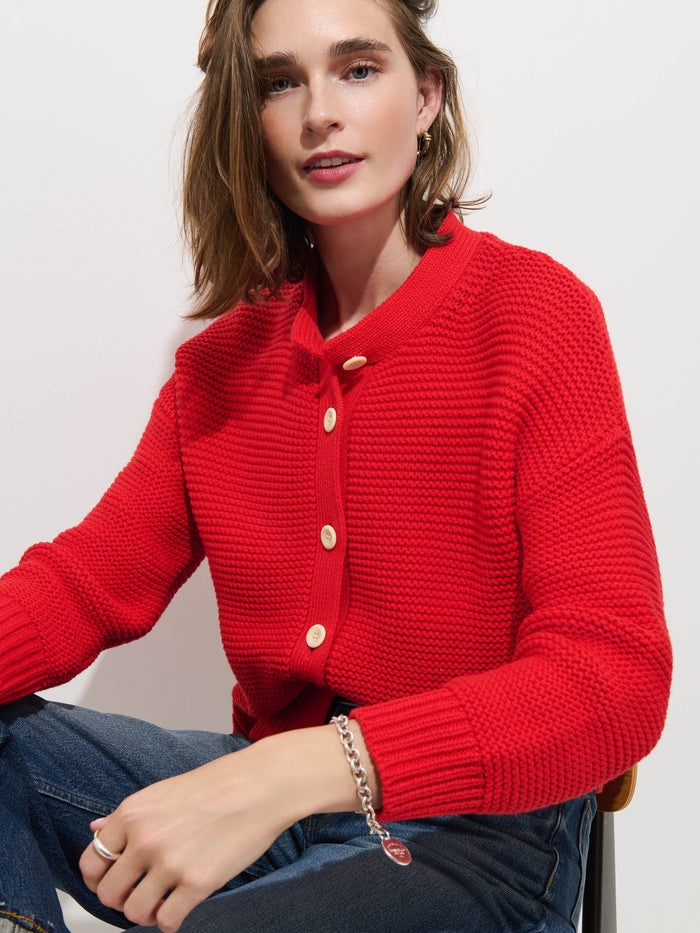 A person sits on a chair wearing the Nico Cardigan in Cotton, a textured red knit sweater with corozo buttons, styled with blue jeans and a silver bracelet, against a plain white background.