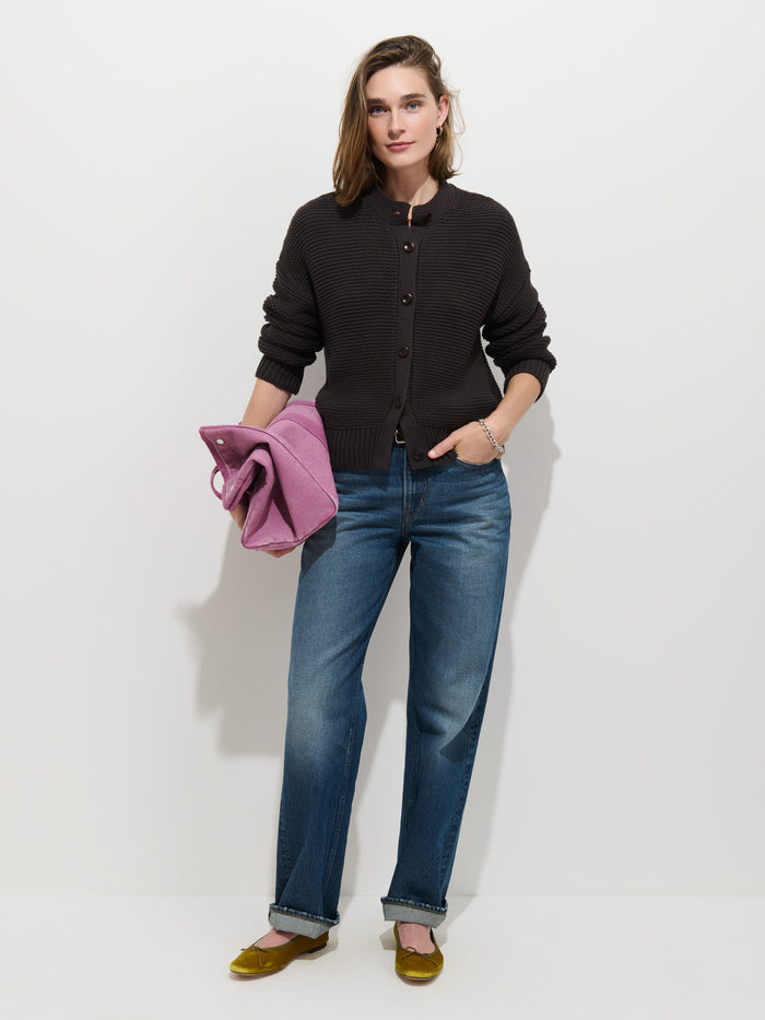 A woman stands against a white background wearing the Nico Cardigan in Cotton, paired with blue jeans and yellow flats. She holds a folded purple coat in her left hand and has her right hand in her pocket.