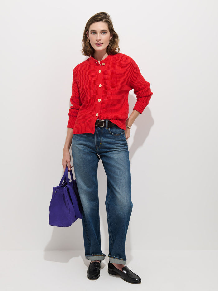 A person stands against a white background, wearing the Nico Cardigan in Cotton—a women's designer red cotton knit sweater with white corozo buttons—paired with blue jeans, a black belt, black loafers, and holding a purple bag.