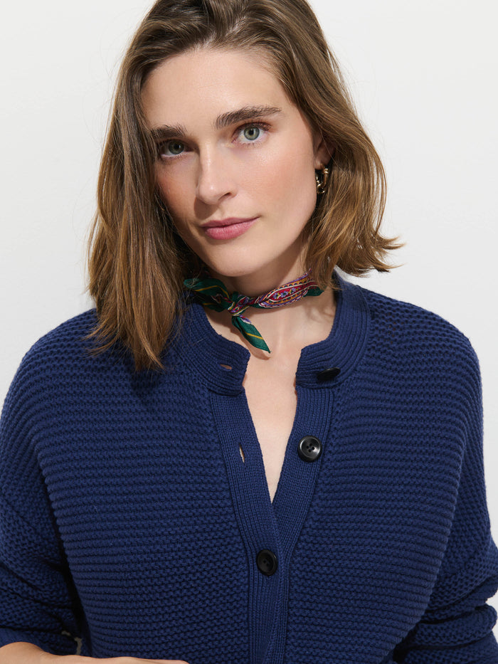 A woman with chin-length brown hair wears the Nico Cardigan in Cotton, a women’s sweater crafted from 100% cotton knit with Corozo buttons down the front, paired with a colorful scarf around her neck, and stands against a plain white background.