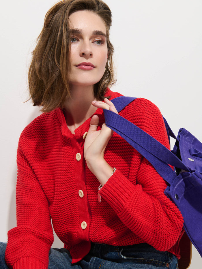 A woman with shoulder-length brown hair wears the Nico Cardigan in Cotton, a textured red knit sweater with corozo buttons, styled with blue jeans and a vibrant purple tote bag over her shoulder. She gazes slightly off-camera with a neutral expression, embodying refined modern style for the contemporary women's designer wardrobe.
