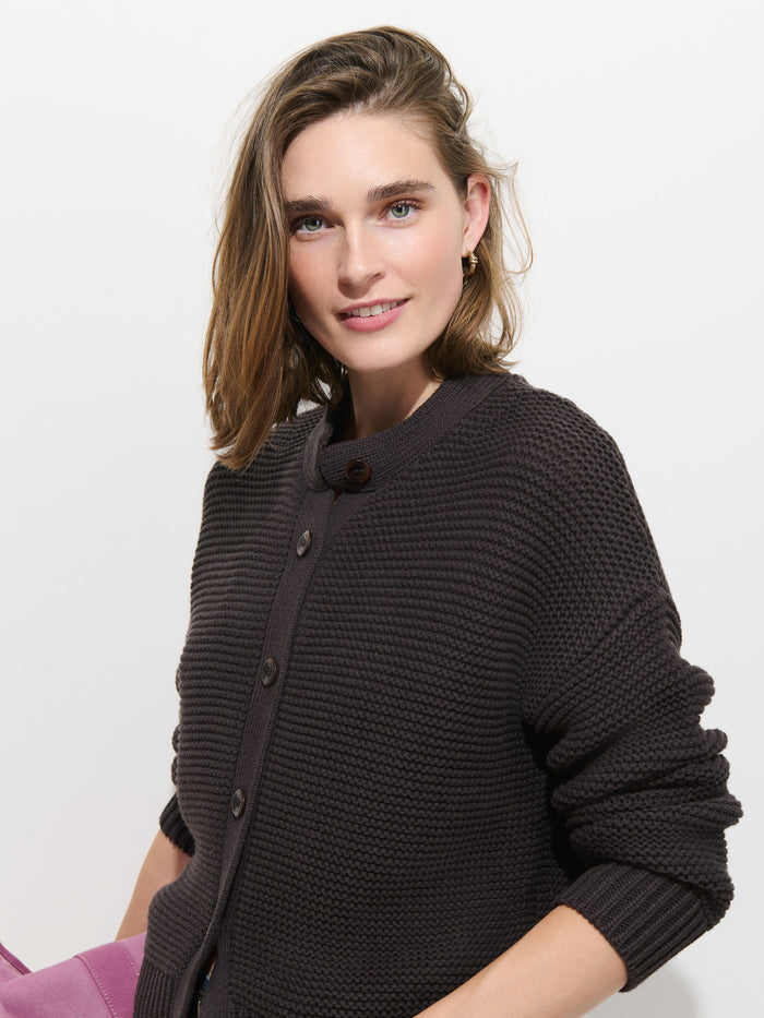 A person with shoulder-length brown hair smiles at the camera against a plain white background, wearing the Nico Cardigan in Cotton, a dark textured women’s designer sweater with corozo buttons. A hint of a pink object is visible in the lower left corner.