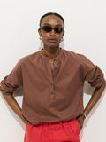 Against a white background, a person wears dark sunglasses, bright red pants, and the Nina Popover in Cotton Voile—an oversized women's designer shirt with rolled-up sleeves and buttons—standing with hands on hips.