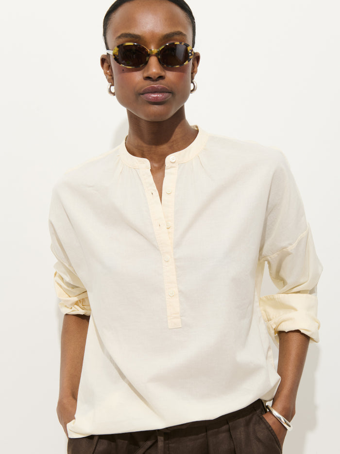 A person in sunglasses, dark pants, and the Nina Popover—an oversized women’s designer shirt in cream 100% cotton voile with rolled sleeves—stands against a plain white background.
