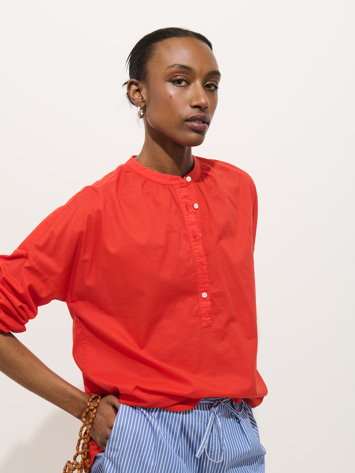 Wearing the Nina Popover in bright red cotton voile with white buttons, paired with blue-and-white striped pants, a person stands against a plain white background holding a brown beaded strap.