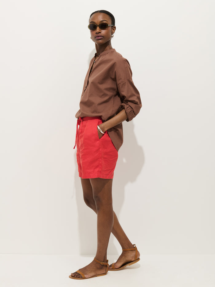A person wearing the Nina Popover in Cotton Voile, round sunglasses, bright red shorts, and tan sandals stands against a plain light background with one hand in their pocket.
