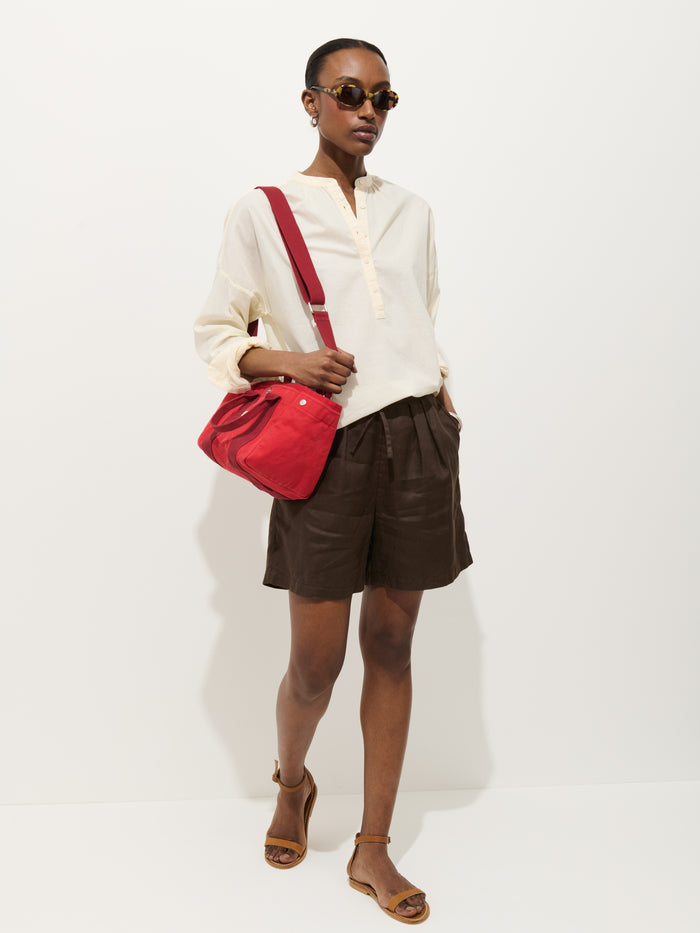 A person wearing sunglasses and the Nina Striped Popover in Cotton Voile stands against a white background, paired with brown shorts, brown sandals, and a red shoulder bag, looking confidently ahead.