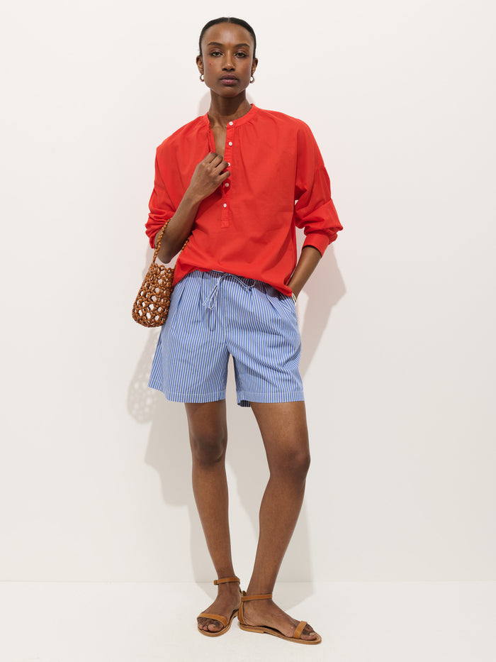 Wearing the Nina Striped Popover in cotton voile, a person stands against a white background paired with blue and white striped shorts, brown sandals, and carrying a small brown woven handbag.