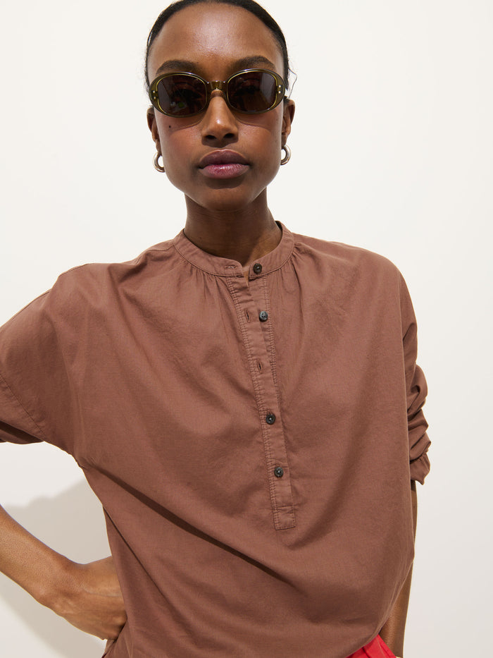 Wearing the Nina Popover in Cotton Voile, paired with dark sunglasses and hoop earrings, a person stands against a plain light background, gazing confidently at the camera.
