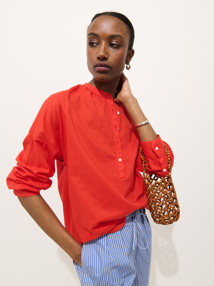 A person poses against a plain background wearing the Nina Striped Popover in Cotton Voile and blue-and-white striped pants, holding a small woven bag and resting one hand on their neck.