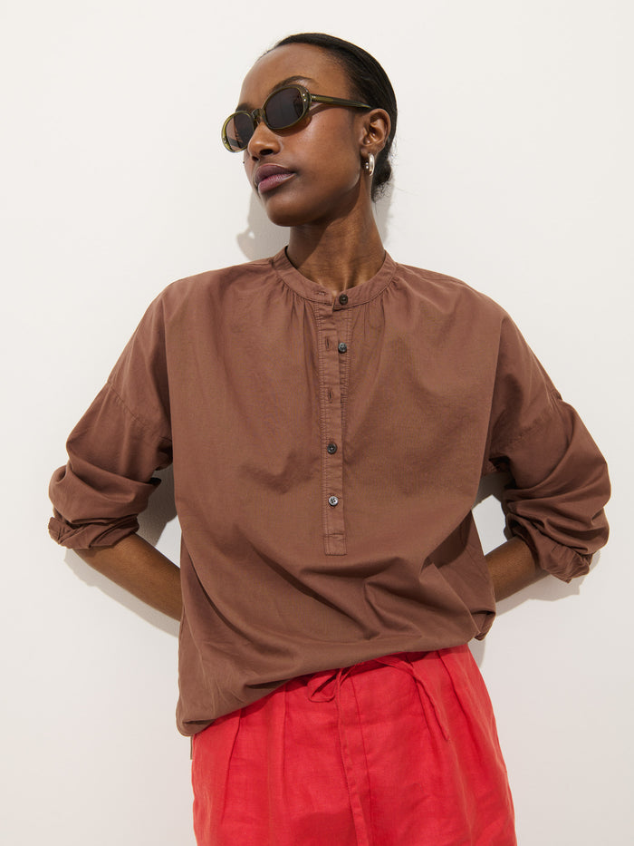 Someone stands against a plain white background with hands behind their back, wearing round sunglasses, red pants, and the Nina Striped Popover In Cotton Voile.
