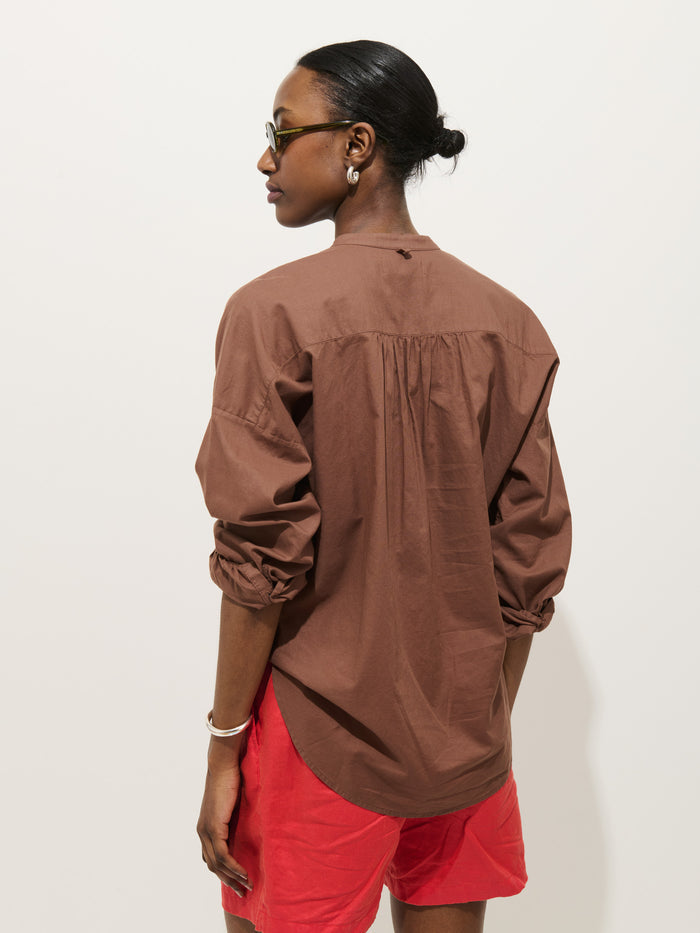 A person with dark hair in a low bun, wearing glasses and the Nina Striped Popover In Cotton Voile, stands facing away from the camera against a plain light background.