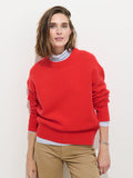 A woman with shoulder-length brown hair wears the Nottinghill Sweater in Wool, a bright red, 100% wool sweater with an oversized fit, layered over a light blue shirt and tan pants. She stands against a plain white background, looking at the camera with a slight smile.