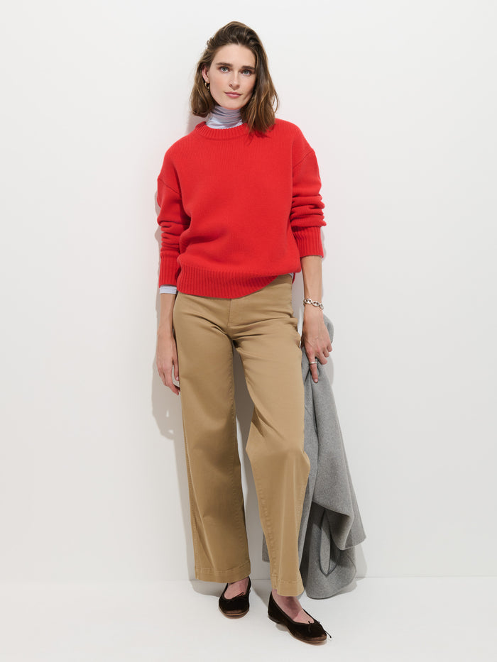 A woman stands against a white background, wearing the Nottinghill Sweater in Wool—a red garment-dyed sweater with an oversized fit—layered over a light blue shirt, paired with beige wide-leg pants and black flats, and holding a gray coat in her left hand.