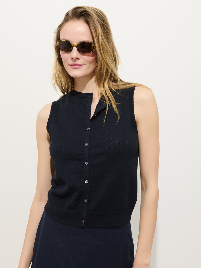 A woman in tortoiseshell sunglasses models the Odette Sweater Tank in Cotton Cashmere, a sleeveless dark navy button-up top, standing against a plain white background and looking off to the side with a relaxed expression.