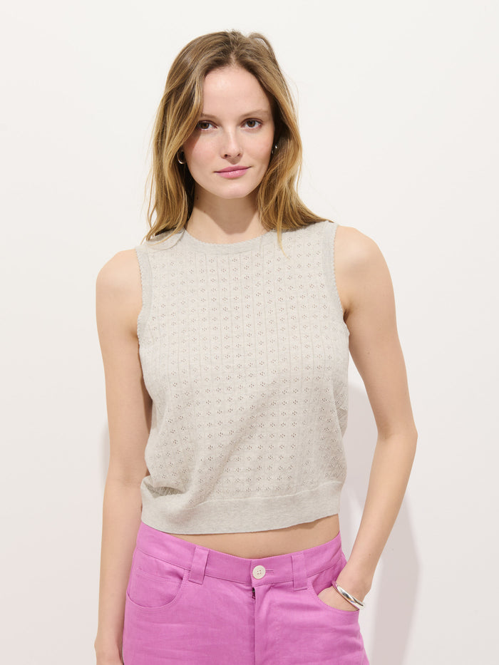 A woman with light brown hair stands against a white background, wearing the Odette Sweater Tank in Cotton Cashmere (beige) and high-waisted pink pants. She has a neutral expression and one hand in her pocket, highlighting the feminine fit.
