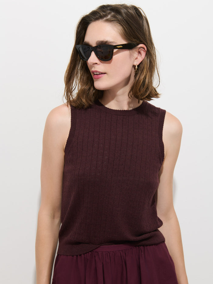A woman with shoulder-length brown hair wears dark sunglasses and the Odette Sweater Tank in Cotton Cashmere—a sleeveless, feminine-fit top in burgundy—paired with a matching skirt, standing against a plain white background.