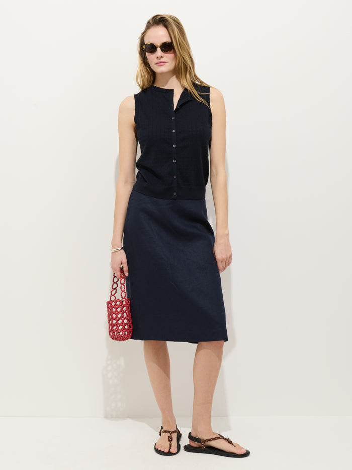 A woman wearing sunglasses, the Odette Sweater Tank in Cotton Cashmere, a dark skirt, black sandals, and holding a red bag stands against a white background.