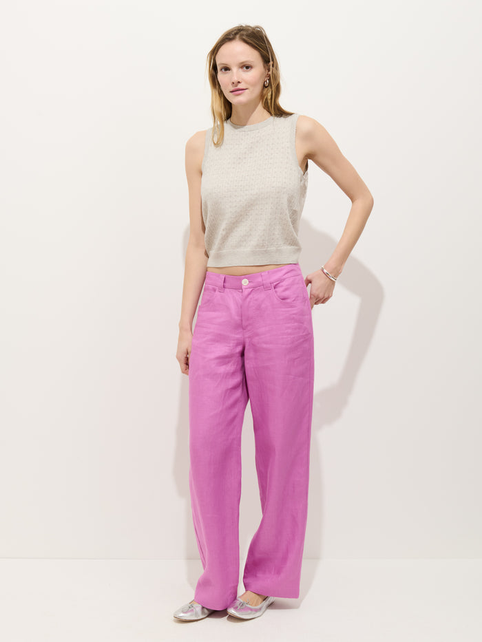 A woman with light hair stands against a white background, wearing the Odette Sweater Tank in Cotton Cashmere—a sleeveless, light gray top with a feminine fit—paired with high-waisted pink wide-leg pants, flats, a bracelet, and earrings.