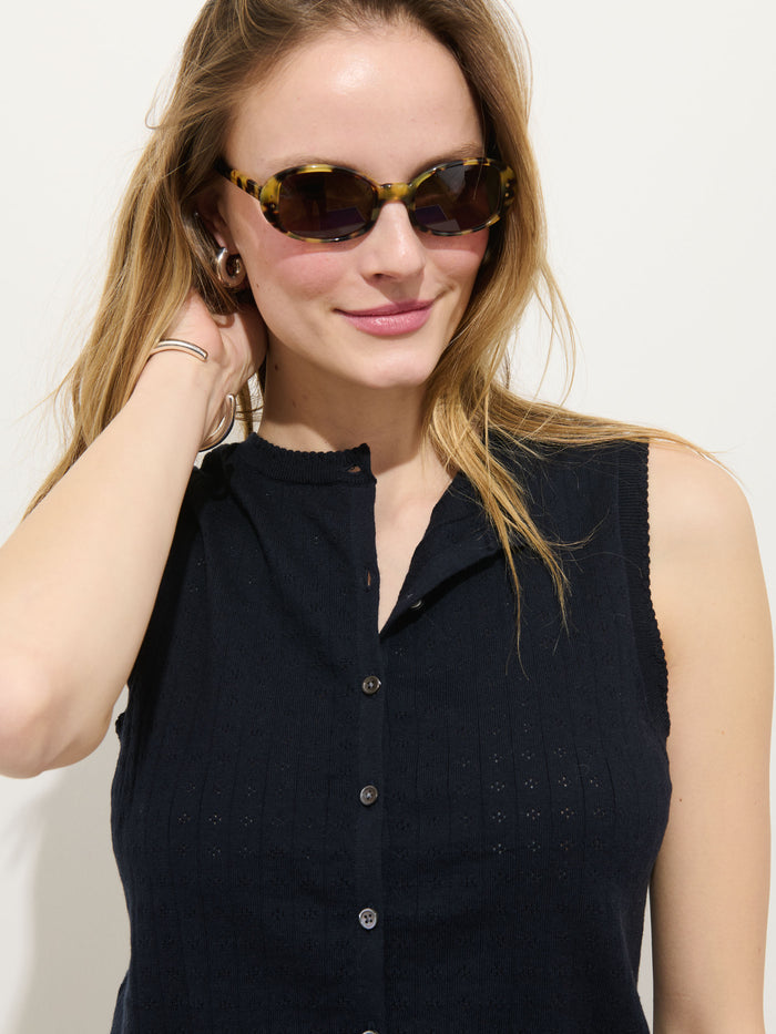 A woman with light brown hair smiles while touching her ear, wearing the Odette Sweater Tank in Cotton Cashmere and tortoiseshell sunglasses, posing against a plain white background.