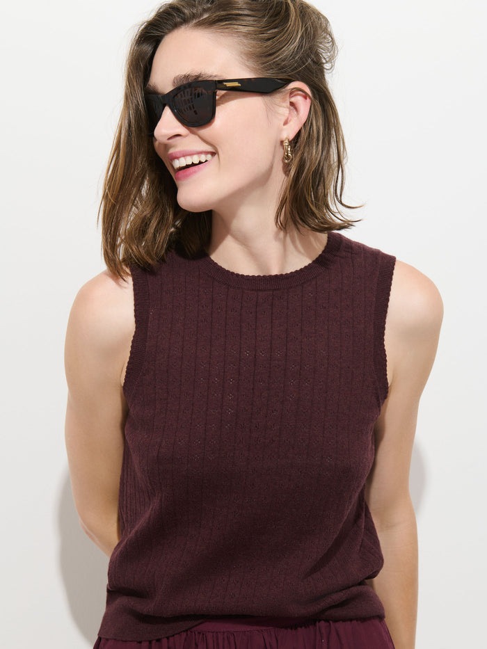 A woman with shoulder-length brown hair wears black sunglasses, gold hoop earrings, and the Odette Sweater Tank in Cotton Cashmere—a sleeveless maroon sweater in a feminine fit—smiling and looking to the side against a plain, light background.