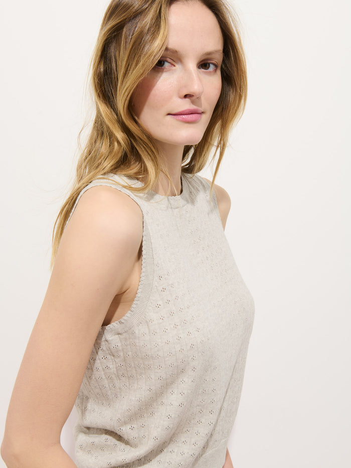 A woman with long, light brown hair models the Odette Sweater Tank in Cotton Cashmere—a sleeveless, light beige top with a feminine fit—standing against a plain background and looking slightly to the side with a subtle smile.