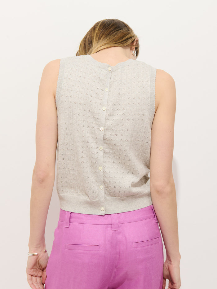 A woman stands facing a white wall, showing the back of her outfit. She wears the Odette Sweater Tank in cotton cashmere, featuring a feminine fit and back buttons, paired with bright pink high-waisted pants. Her hair is pulled to the side.