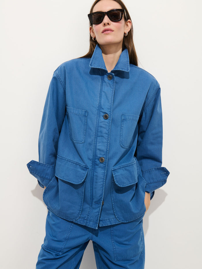 A person wearing dark sunglasses and the Painter Jacket In Herringbone—an oversized blue cotton jacket with large front pockets—plus matching blue pants, stands against a plain white background with hands in pockets.