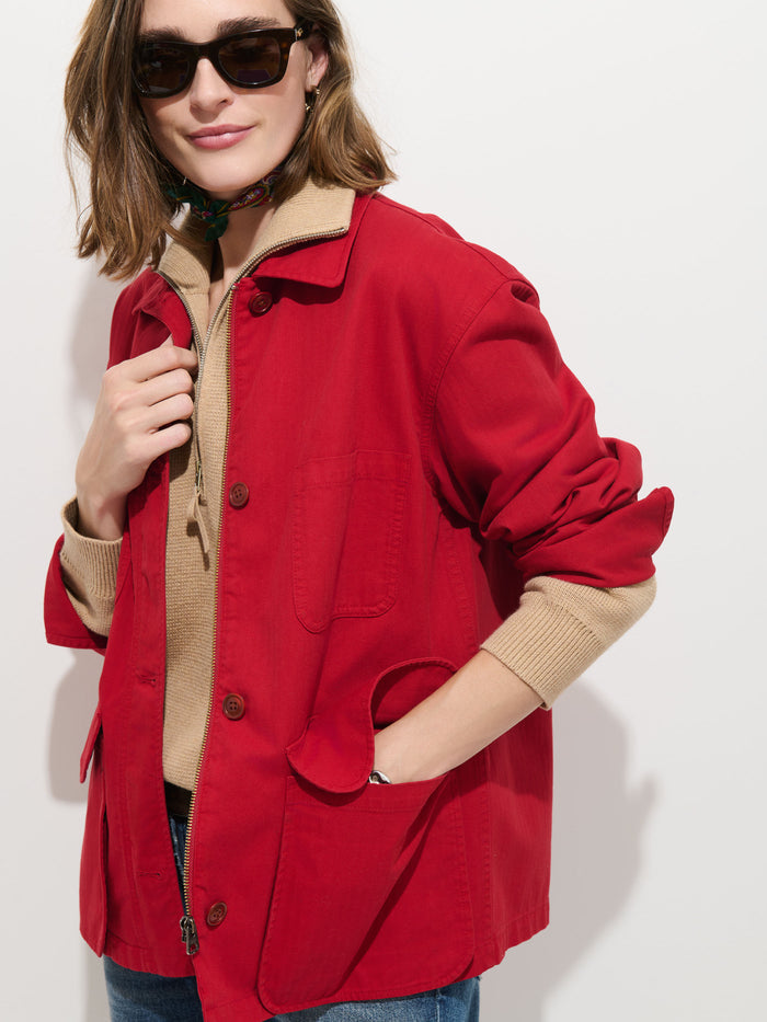 A woman wearing sunglasses stands against a white background, dressed in the Painter Jacket In Herringbone—a red, oversized-fit cotton herringbone jacket—layered over a tan sweater with blue jeans. One hand is tucked in her pocket while the other touches her jacket collar.