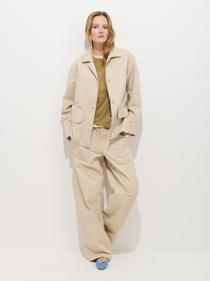 A person stands against a white wall in an oversized Painter Jacket In Herringbone and wide-leg trousers, with a tan shirt and light blue shoes. Their hands are in their pockets and they appear relaxed.