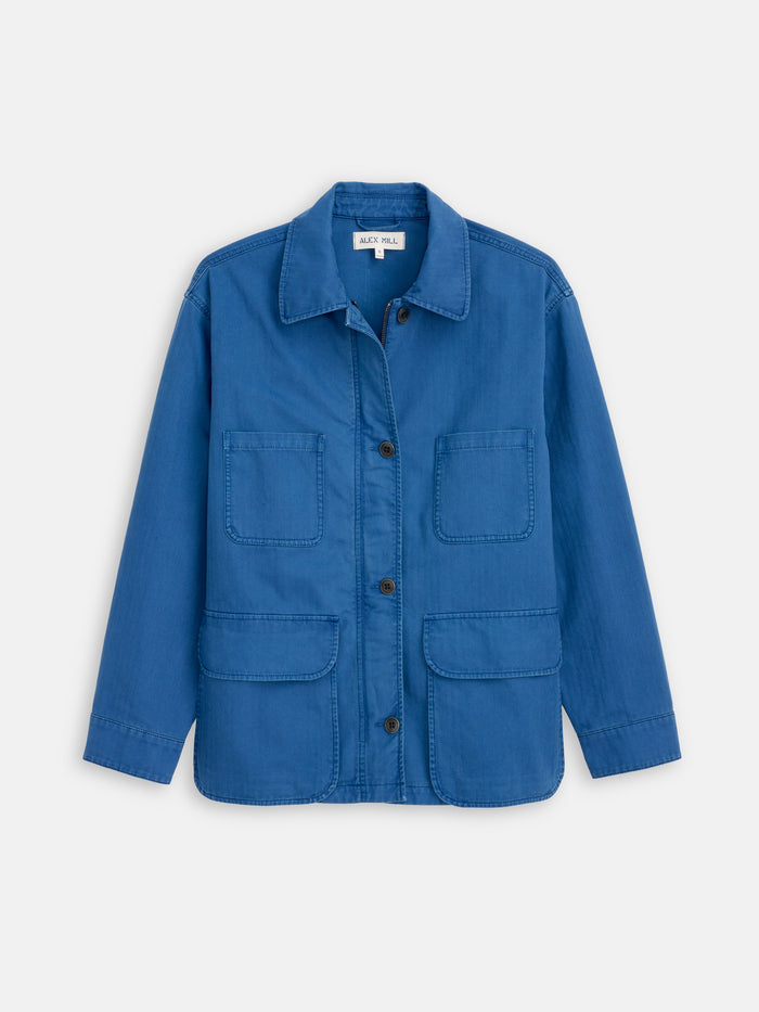 The Painter Jacket In Herringbone is a blue cotton jacket with a collar, long sleeves, and four front patch pockets, made from durable herringbone fabric and shown on a plain white background.